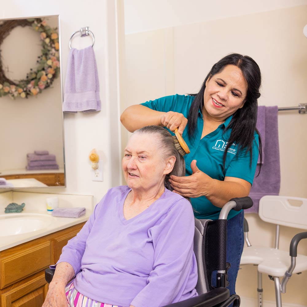 A female caregiver wearing a mask stands behind male patient while brushing his hair. A female caregiver stands behind a female patient while brushing her hair.